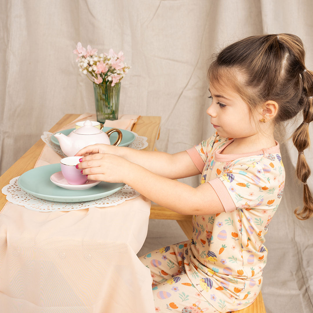 toddler girl sits at a table. she is preparing for a tea party. there are tea cups, plate, and flowers on the table. she wears the "egg hunt" matching pajama set. the "egg hunt" print is a easter theme. the colors are pastel. you can find flowers, carrots, easter bunnies in easter eggs, and chicks who have half an easter egg on their head. 