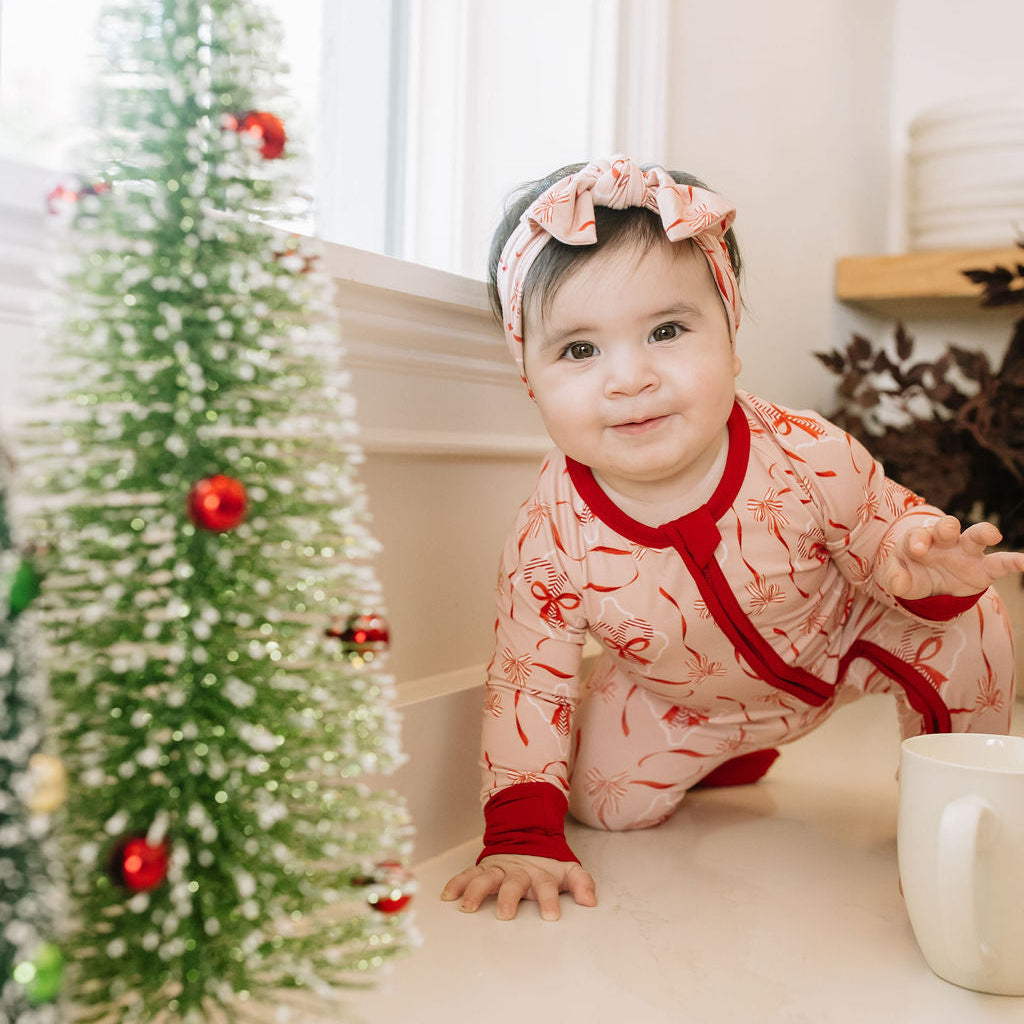 Baby wearing Emerson and Friends Candy Cane Coquette bamboo Christmas pajamas with a matching pink bow headband, crawling beside a decorated Christmas tree with red ornaments.