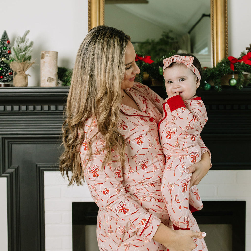 Mom holding her baby in matching Emerson and Friends Candy Cane Coquette Christmas bamboo pajamas, featuring a pink and red bow print, in front of a festive holiday fireplace.