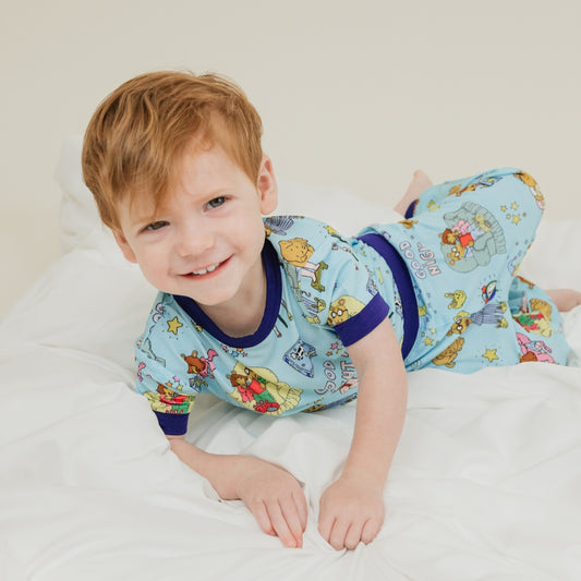 A sweet moment of a child smiling while relaxing in a bean bag, wearing our PBS Good Night Arthur™ collaboration featuring pajamas adorned with Arthur and friends in a cozy bedtime scene complete with storybooks, toothbrushes, bunny slippers, a sleeping puppy, crescent moons with sleep caps, twinkling stars, and the words "GOOD NIGHT!" on a soft blue background.