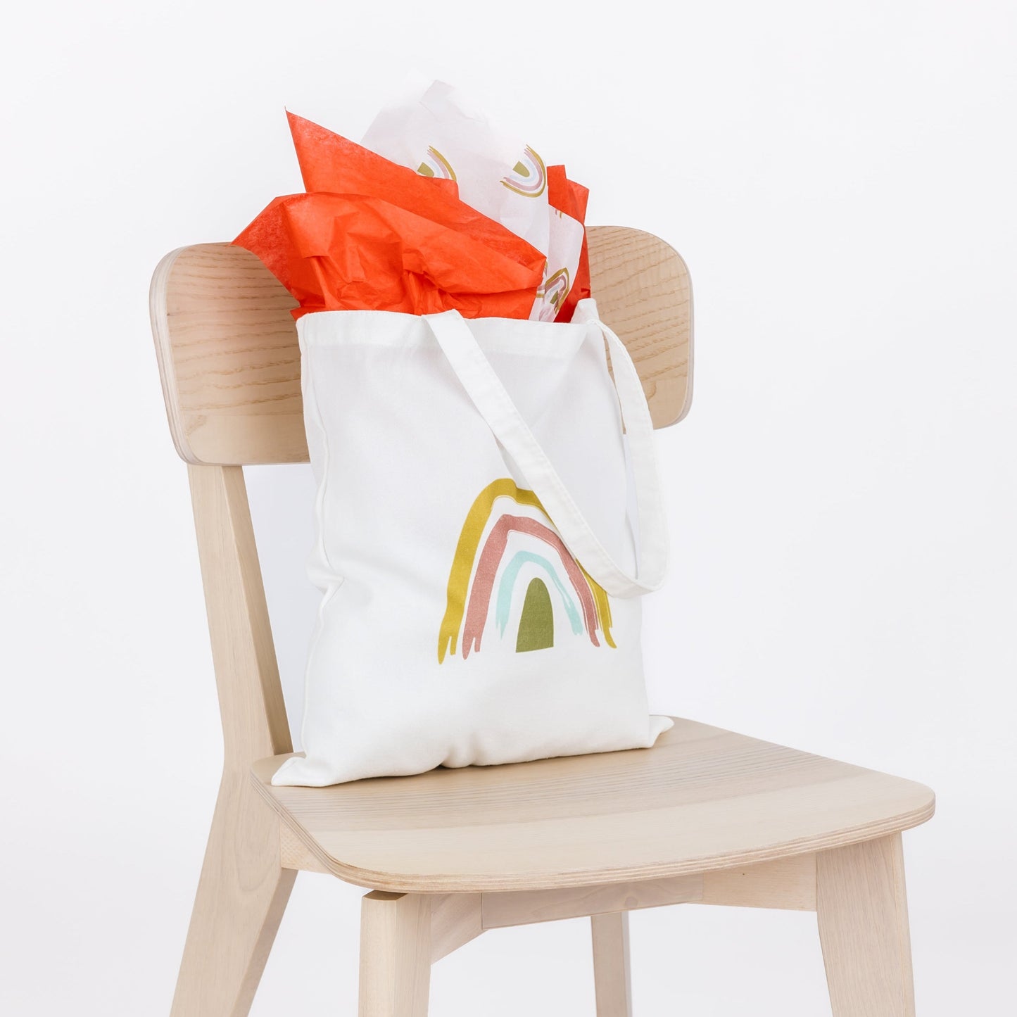 White tote bag with rainbow design on a wooden chair against a white background