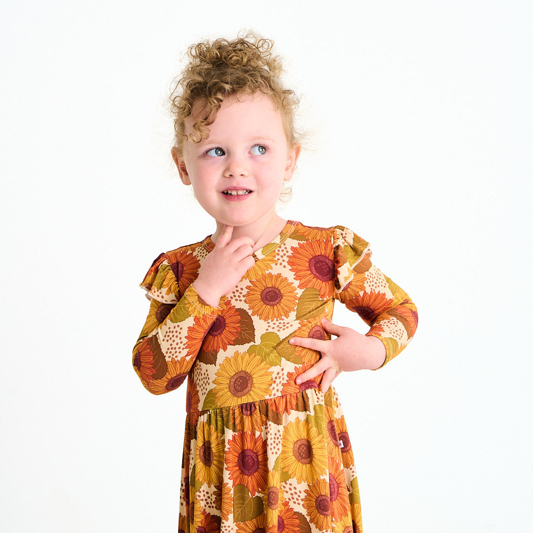 young girl wears the "sunflower" twirl dress. the "sunflowers" print is a sunflower floral design. it has a variety of yellow, orange, and red sunflowers, mixed into green leaves. 