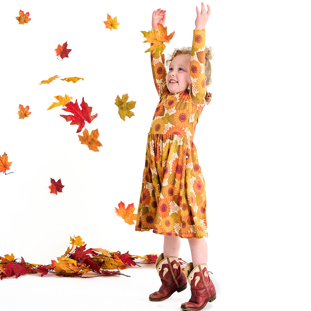 little girl throws fall leaves into the air. you can see her in the "sunflower" twirl dress, with some cowgirl boots on. the "sunflowers" print is a sunflower floral design. it has a variety of yellow, orange, and red sunflowers, mixed into green leaves. 