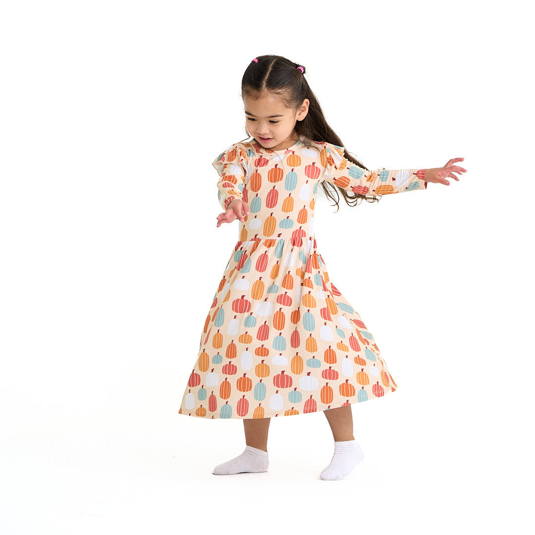 girl is captured twirling in her "pumpkin patch" twirl dress. the 'pumpkin patch" print is a mix of boho styled and colored pumpkins. you can see white, blue, orange, and red pumpkins in.different shapes and sizes. this is all on a pastel orange background. 