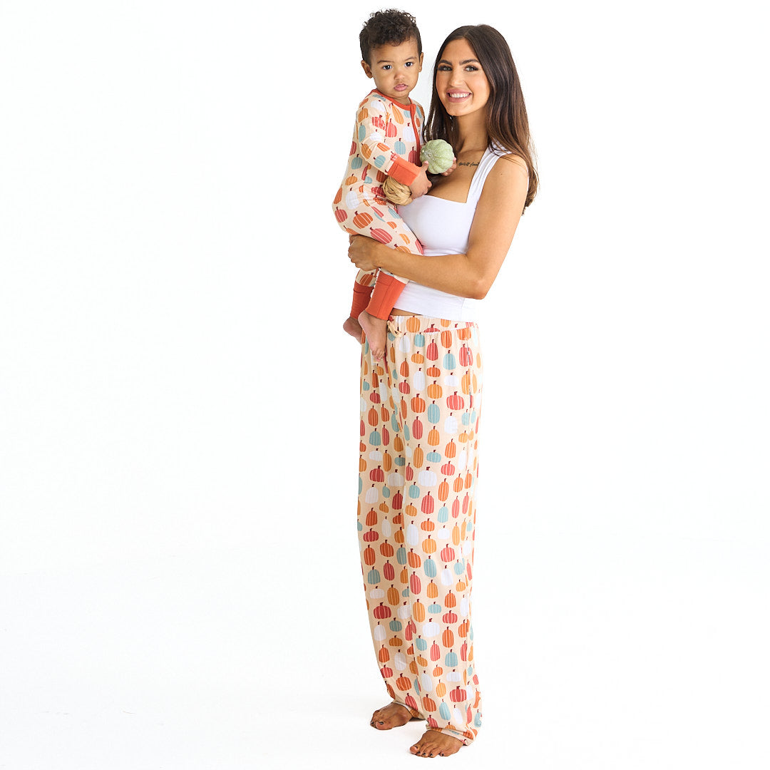 mom smiles and holds her son on her hip. they both wear "pumpkin patch" apparel. mom is in the relaxed pants and the son is in the convertible. the 'pumpkin patch" print is a mix of boho styled and colored pumpkins. you can see white, blue, orange, and red pumpkins in.different shapes and sizes. this is all on a pastel orange background. 