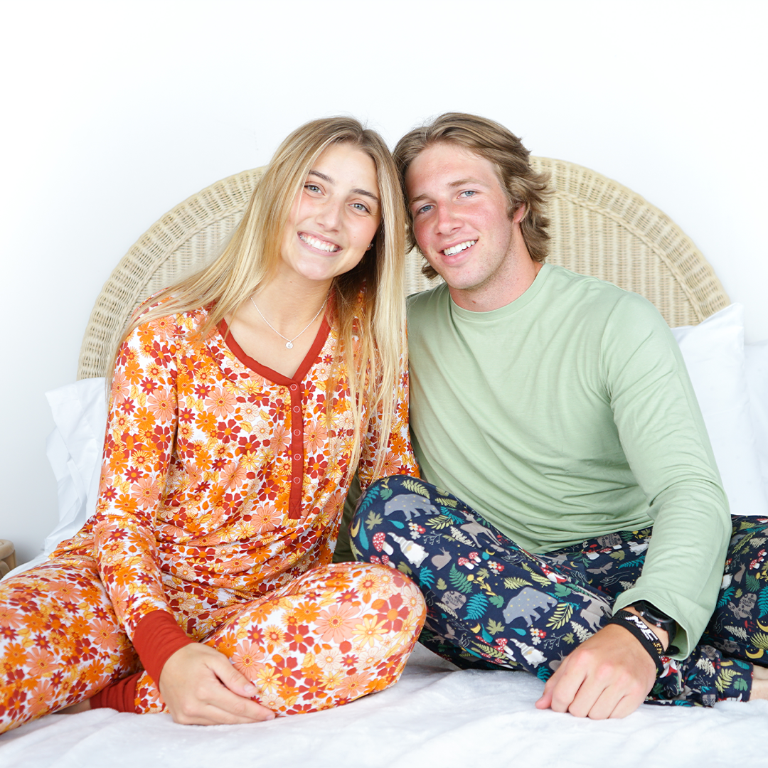 a couple sits on a white bed and smile with their heads together. the girl on the left wears the "fall floral" women's top and joggers. the boy on the right wears the "prehistoric friends" relaxed pants. the "fall floral" print is a floral flowered print that is filled with fall colors and flowers. 