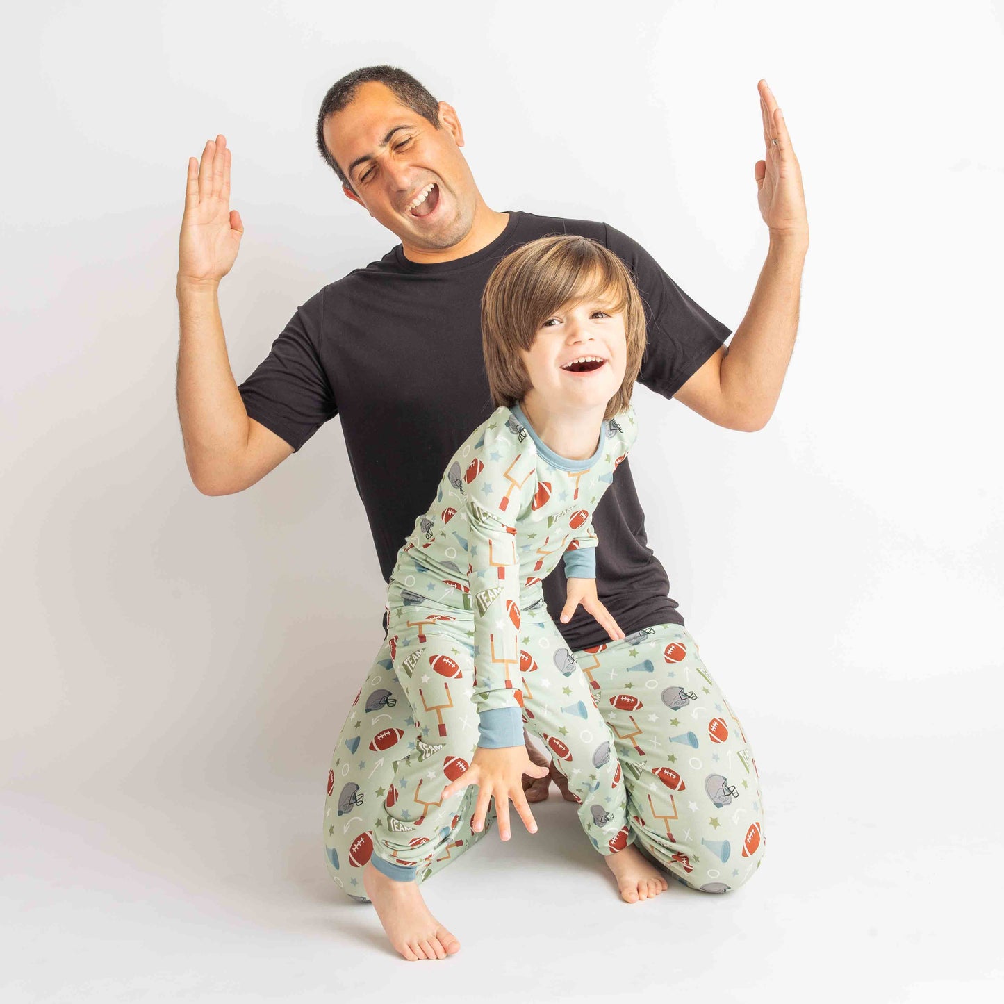 Dad and son laughing while wearing long-sleeve toddler set, and lounge pants in the green football print called "Game Day" Featuring footballs, goal posts, stars and more!