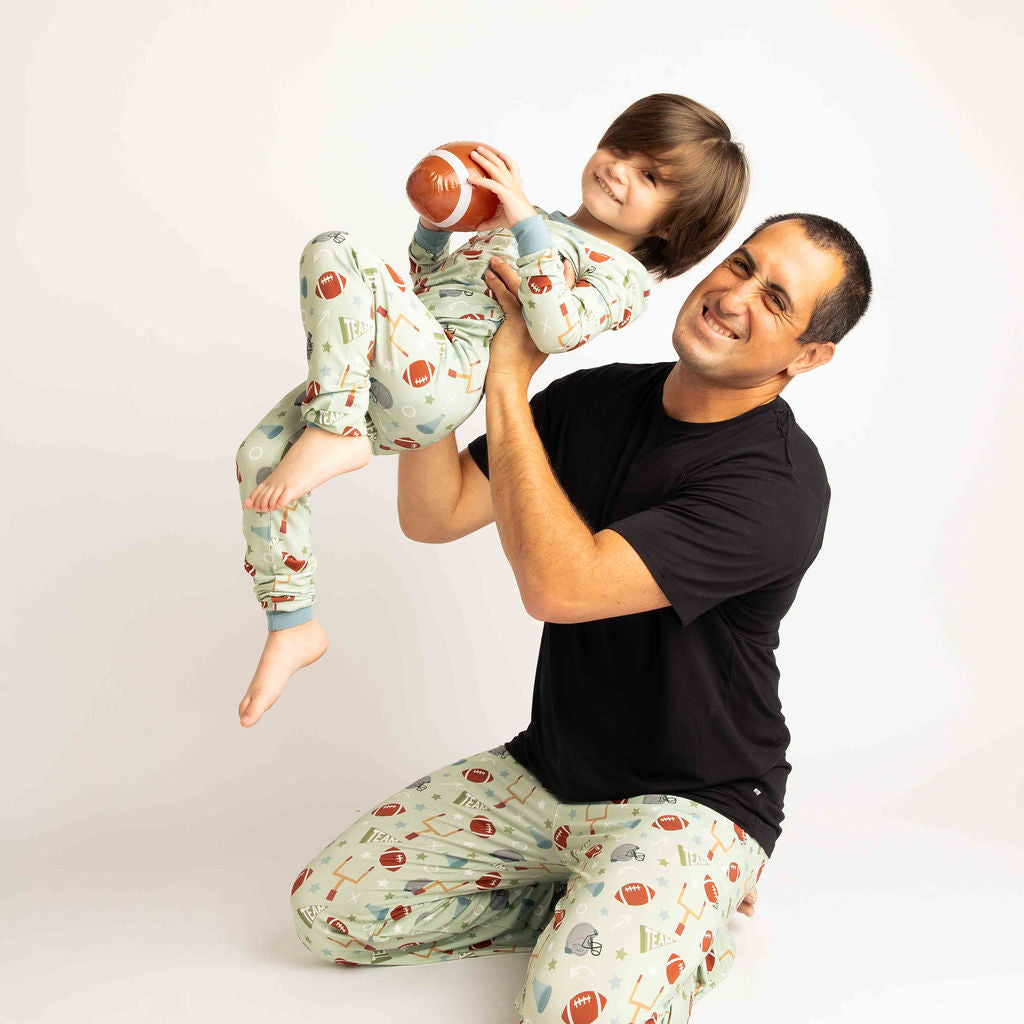 Dad holding son up in the aire with football in his hand. Both wearing green football print called "Game Day" Featuring footballs, goal posts, stars and more! 