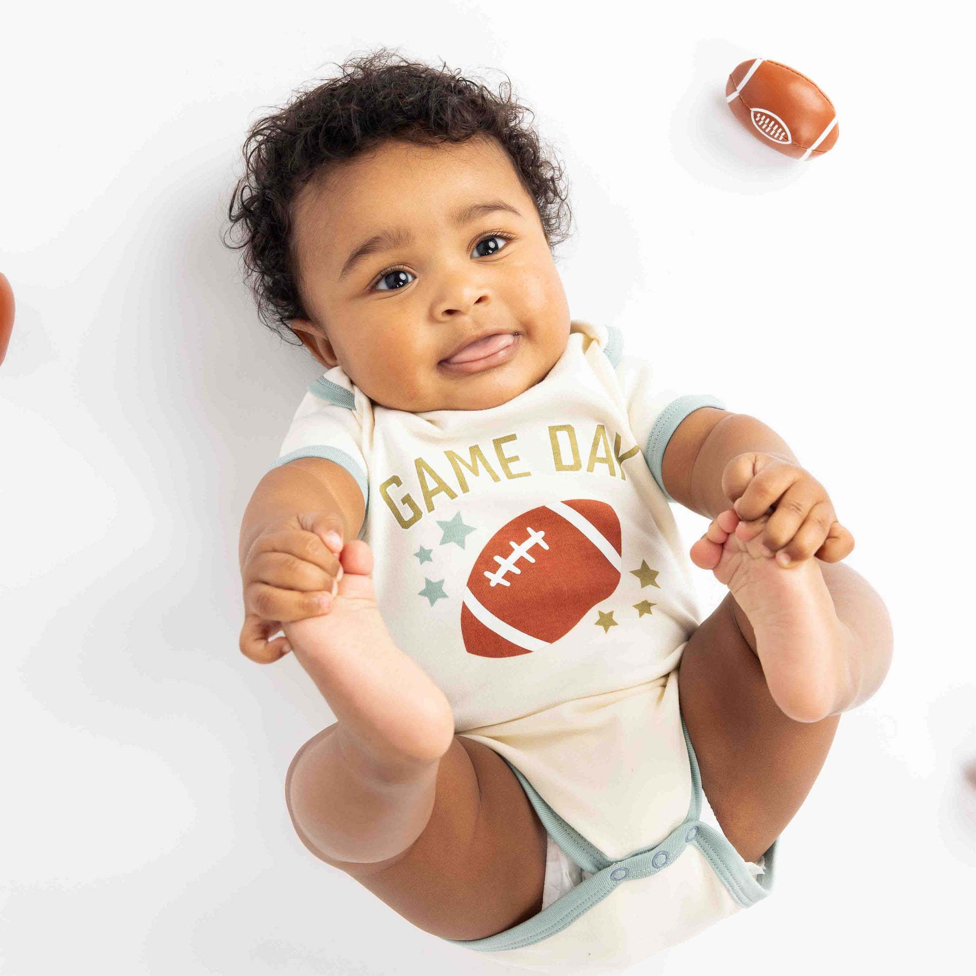 baby laying down with footballs around him. Modeling a blue ringer white onesie with a football, stars and the phrase "game Day" on it. 