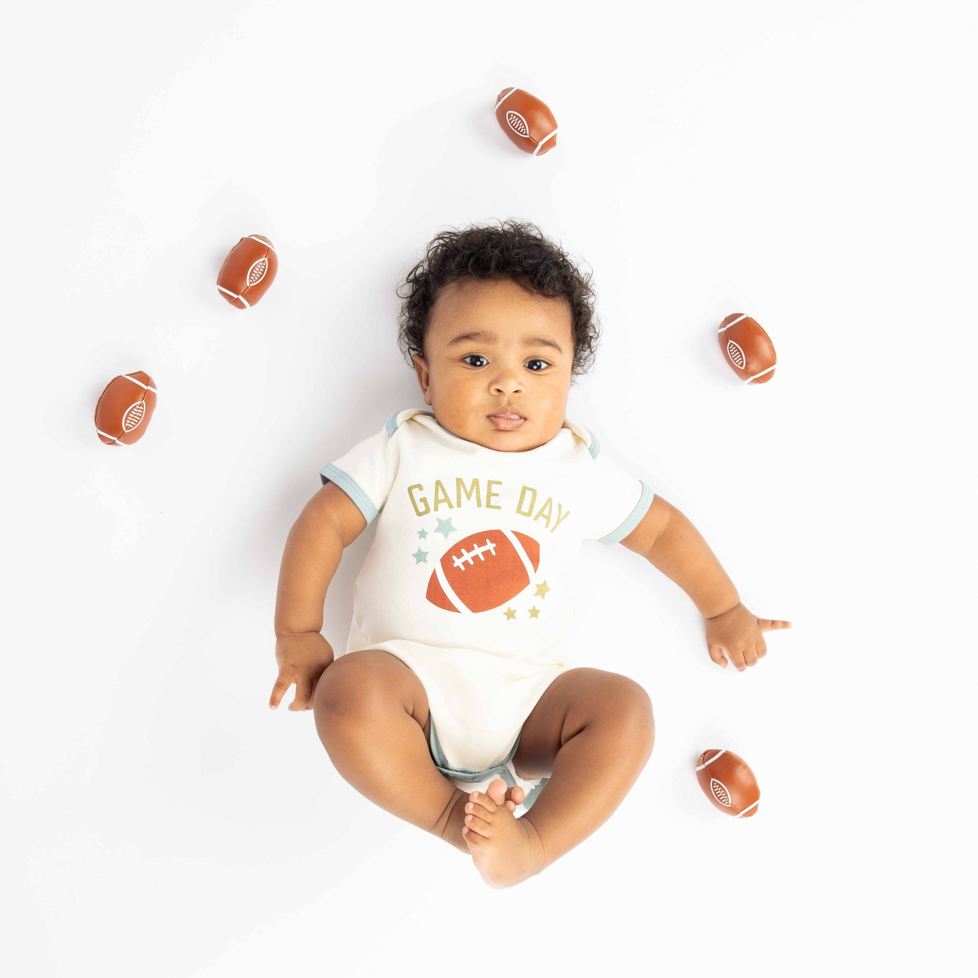 baby laying down with footballs around him. Modeling a blue ringer white onesie with a football, stars and the phrase "game Day" on it. 