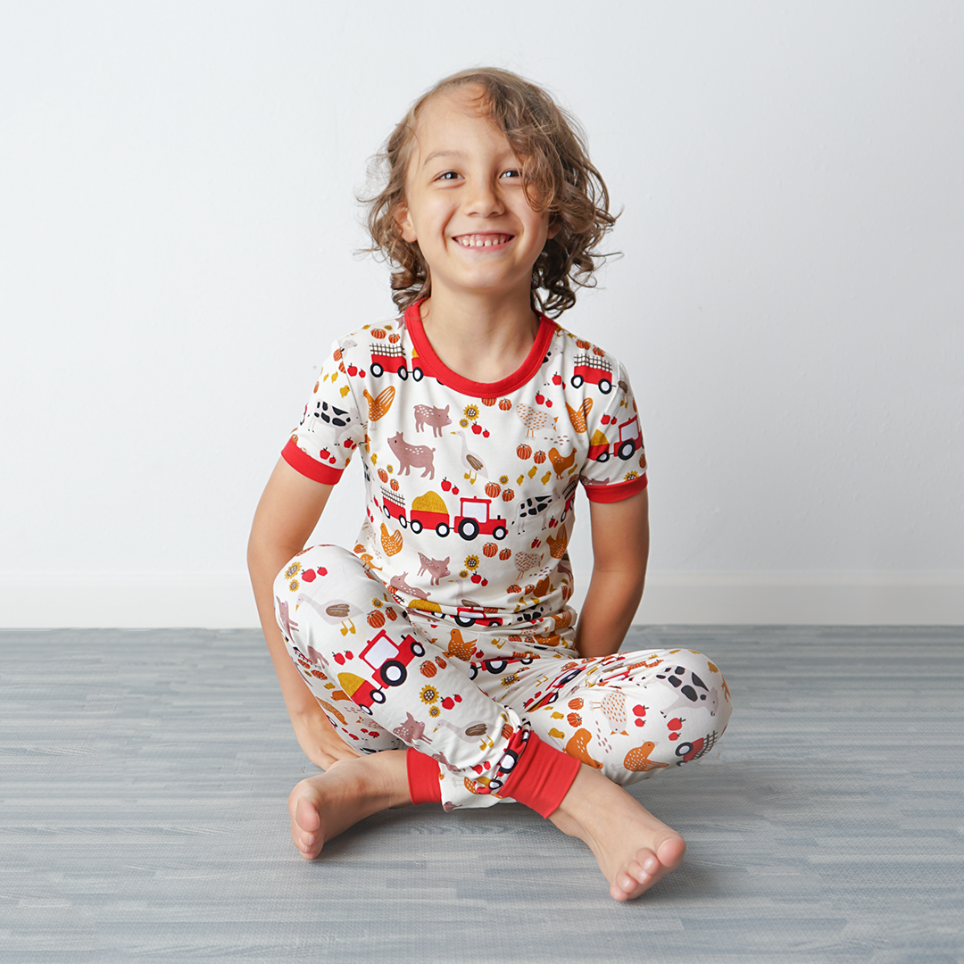 a young girl sits happily on the floor. she is wearing the "farm friends" matching pajama set. the "farm friends" print is a collection of chickens, roosters, pigs, bails of hay, tractors, chicks, apples, and pumpkins. it's a white background and flashed of reds, yellows, and oranges, to bring out the farm aesthetic.