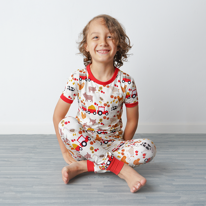 a young girl sits happily on the floor. she is wearing the "farm friends" matching pajama set. the "farm friends" print is a collection of chickens, roosters, pigs, bails of hay, tractors, chicks, apples, and pumpkins. it's a white background and flashed of reds, yellows, and oranges, to bring out the farm aesthetic.