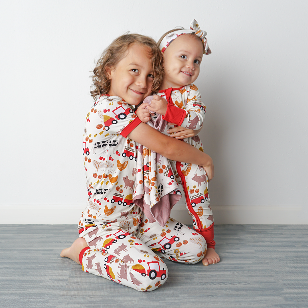 2 sisters sit on the floor hugging one another. all of them in "farm friends" attire. the oldest is in the 2-piece matching pajama set. the youngest is in the convertible and the headband. she holds the lovey. the "farm friends" print is a collection of chickens, roosters, pigs, bails of hay, tractors, chicks, apples, and pumpkins. it's a white background and flashed of reds, yellows, and oranges, to bring out the farm aesthetic.