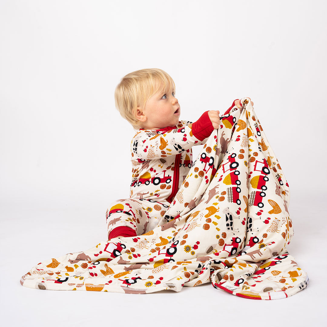 a small baby wears the 'farm friends" convertible and holds up the "farm friends" bamboo blanket. the "farm friends" print is a collection of chickens, roosters, pigs, bails of hay, tractors, chicks, apples, and pumpkins. it's a white background and flashed of reds, yellows, and oranges, to bring out the farm aesthetic.