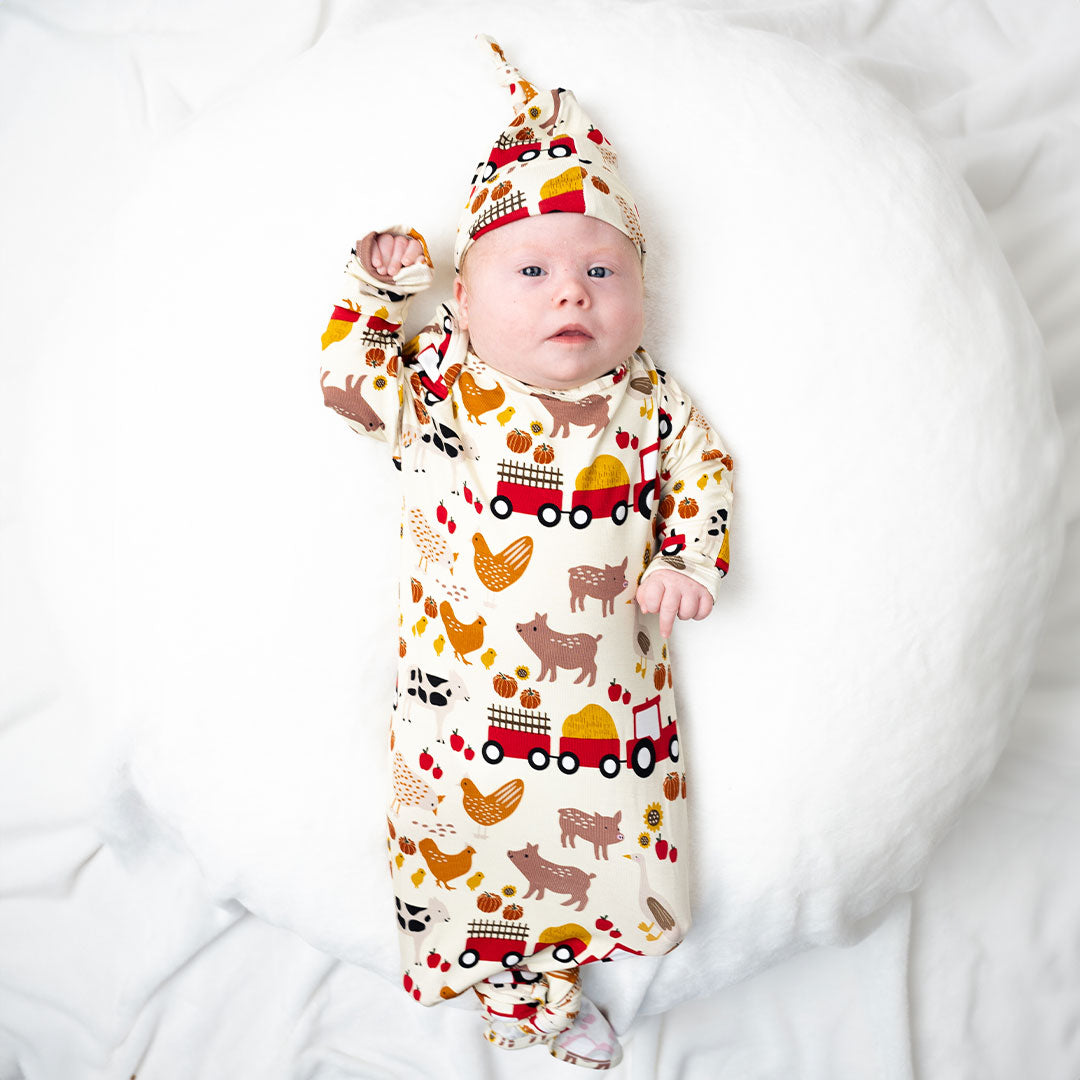 a baby lays on a white pillow. the baby wears the "farm friends" gown and hat set. the gown is tied in a knot at the bottom. the "farm friends" print is a collection of chickens, roosters, pigs, bails of hay, tractors, chicks, apples, and pumpkins. it's a white background and flashed of reds, yellows, and oranges, to bring out the farm aesthetic.