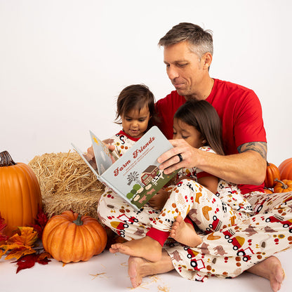 father and 2 daughters sit next to some hay and pumpkins. they read the book "farm friends"