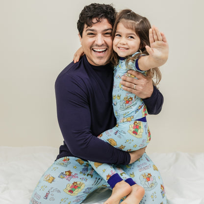 A heartwarming moment captures the father with daughter smiling, with them wearing our PBS Good Night Arthur™ collaboration featuring pajamas adorned with Arthur and friends in a cozy bedtime scene complete with storybooks, toothbrushes, bunny slippers, a sleeping puppy, crescent moons with sleep caps, twinkling stars, and the words "GOOD NIGHT!" on a soft blue background.