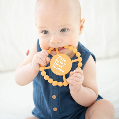 little baby holding a Round orange baby teether that says this is my first rodeo on the front