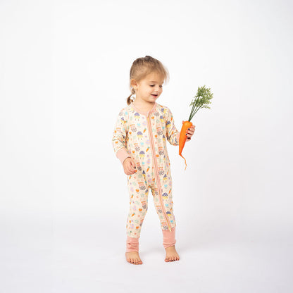 toddler girl holds a large carrot and wears the "egg hunt" convertible. the "egg hunt" print is a easter theme. the colors are pastel. you can find flowers, carrots, easter bunnies in easter eggs, and chicks who have half an easter egg on their head. 