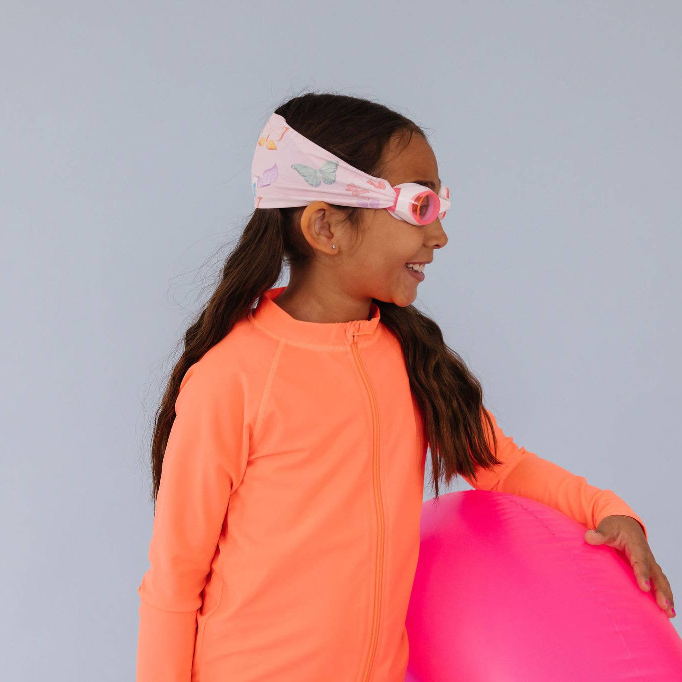 Young girl in an orange shirt holding a pink ball against a gray background with Splash Place flutter swim goggles with butterfly print fabric strap
