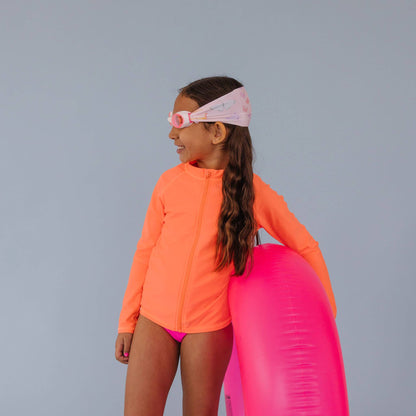 Child in orange long-sleeve shirt and pink swimsuit holding a pink inflatable tube against a gray background wearing Splash Place flutter swim goggles with butterfly print fabric strap