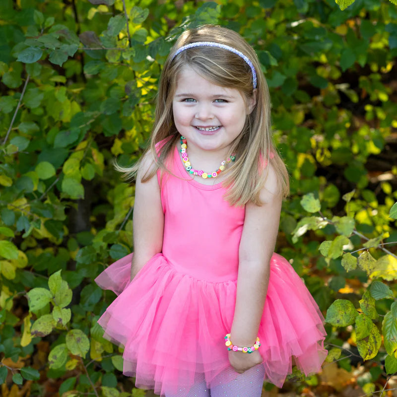 a little girl in a pink dress standing outside in front of some bushes wearing a purple headband and a fruit themed necklace
