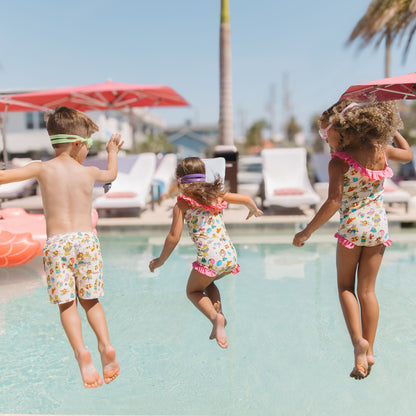 Siblings jumping into the pool in our Arthur's Family Vacation one piece swimwear featuring vibrant pink trim and the boys swim trunks with a playful print of Arthur™ in swimwear surrounded by fun beach props — perfect for family getaways.