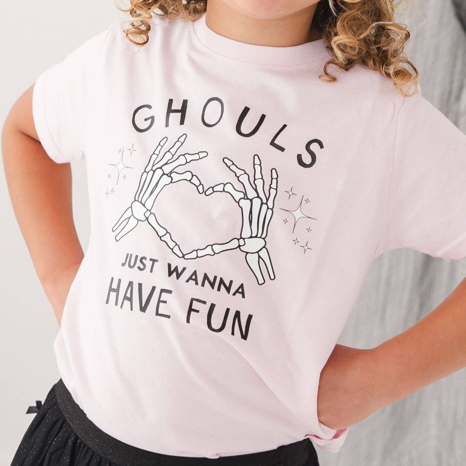 A child wearing a light pink short-sleeve T-shirt with a black graphic of skeleton hands forming a heart and the text 'GHOULS JUST WANNA HAVE FUN'.