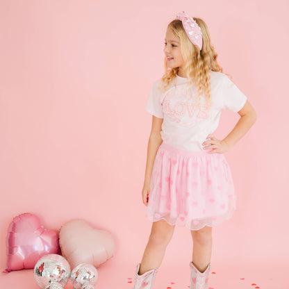 Young girl in a white t-shirt and pink tutu skirt standing against a pink background with heart-shaped balloons.