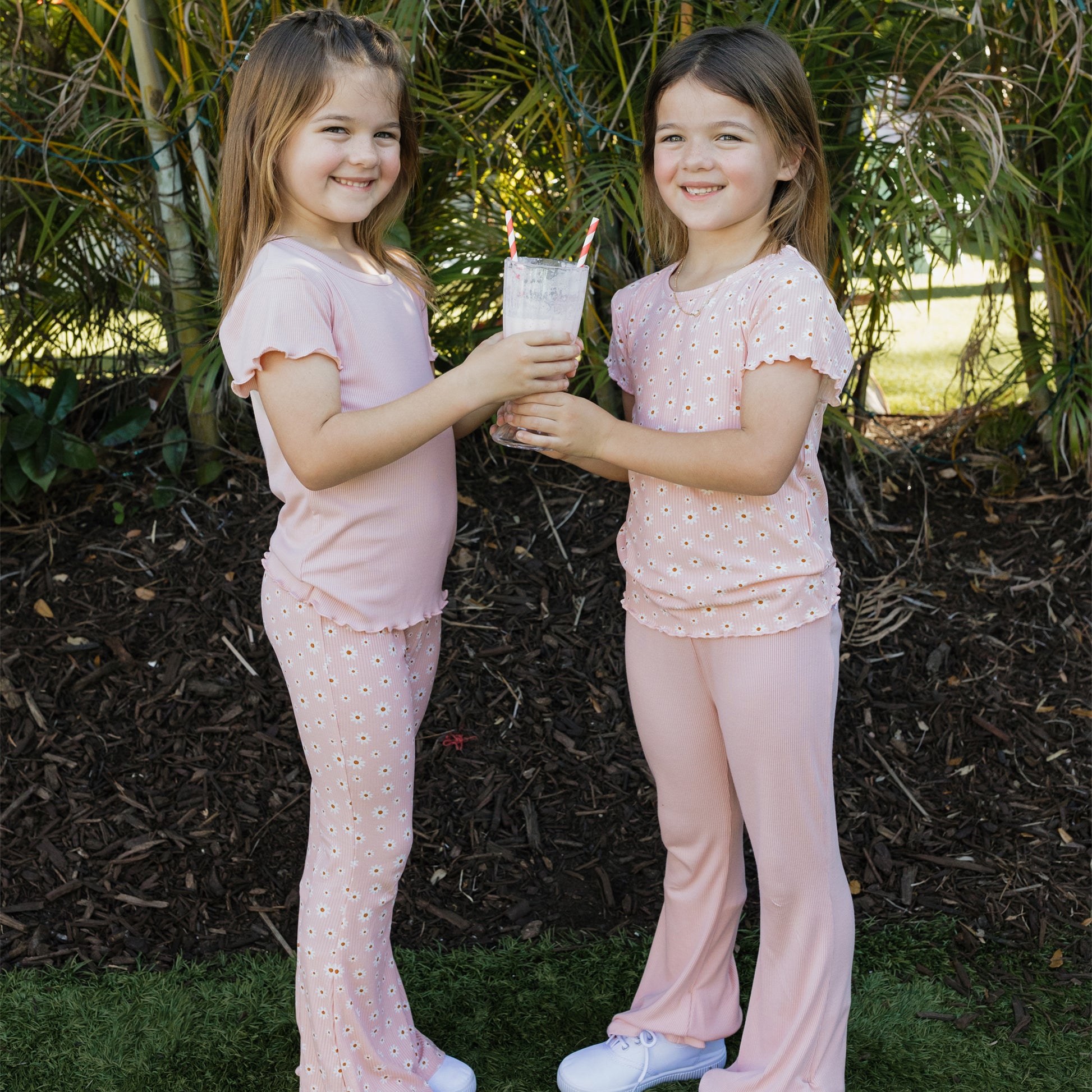 Two young girls in matching pink outfits holding a glass with straws outdoors.