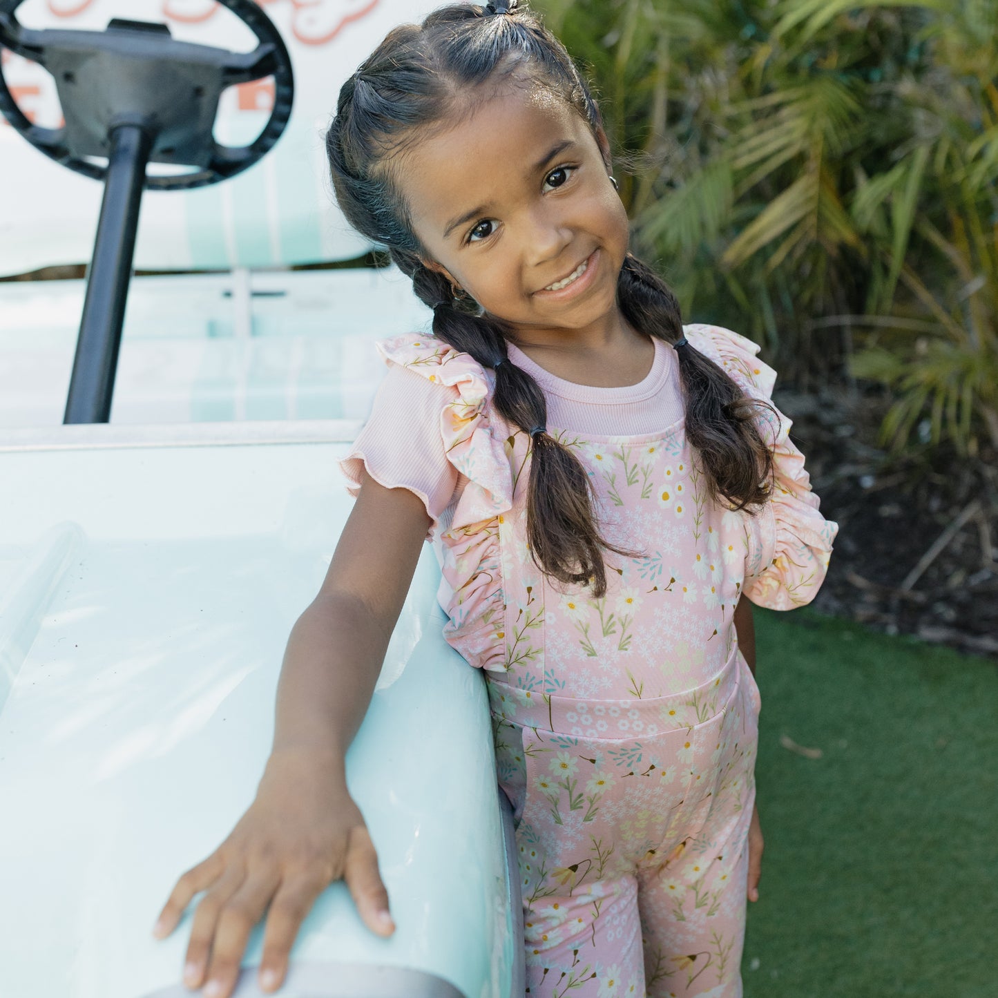Young girl in a pink floral outfit standing next to a golf cart with greenery in the background