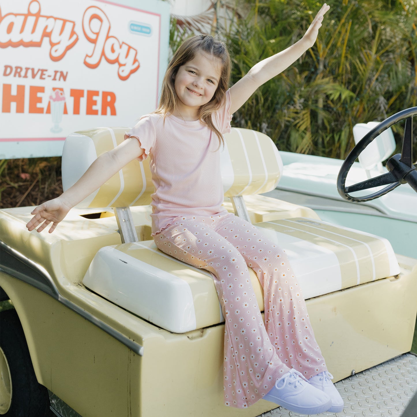 Young girl sitting on a vintage car with an open arms pose, in front of a Dairy Joy Drive-In Theater sign.