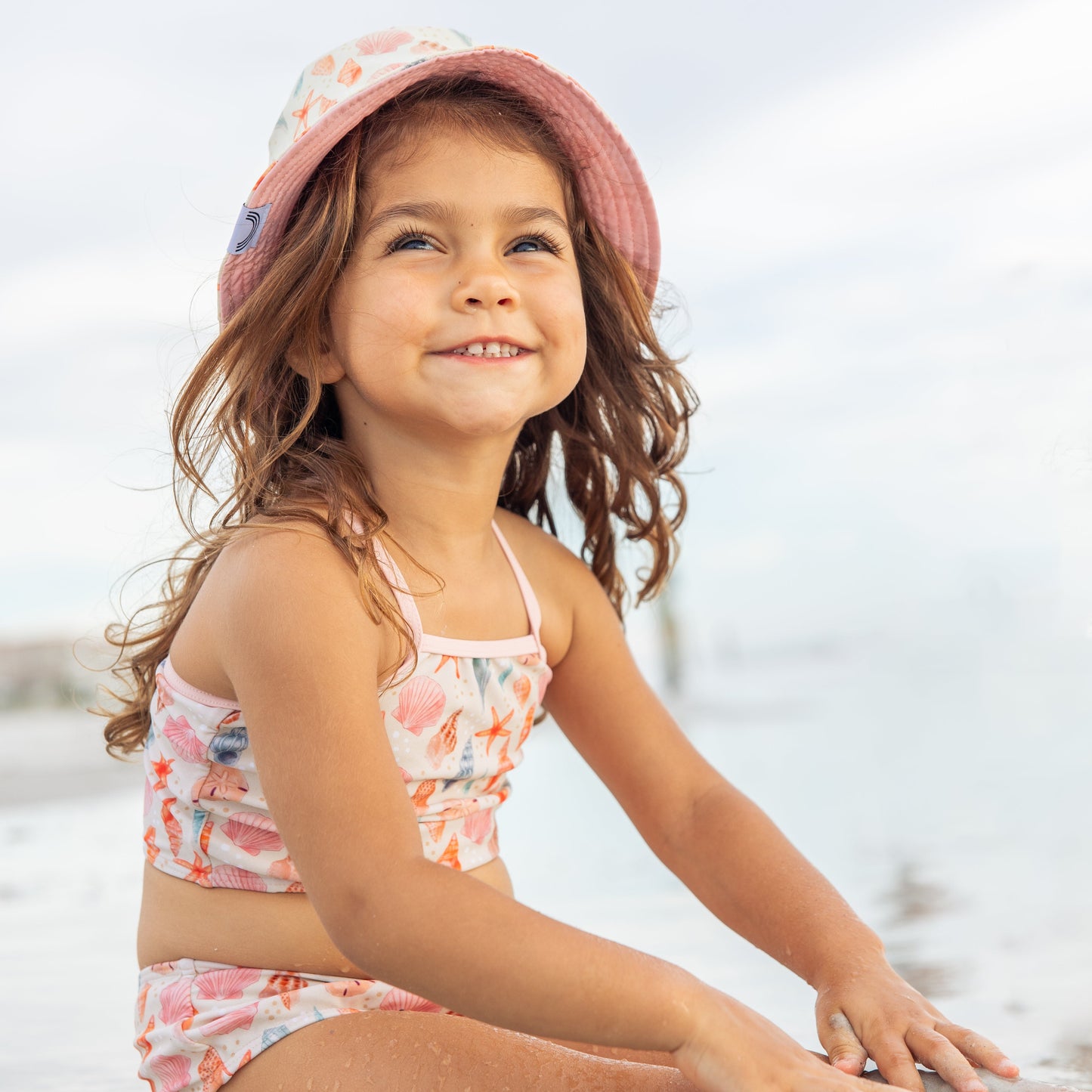 Little girl in the sand wearing the Sandy Seashells Bikini Girls Swimsuit and matching Sandy Seashells Reversible Bucket Hat.