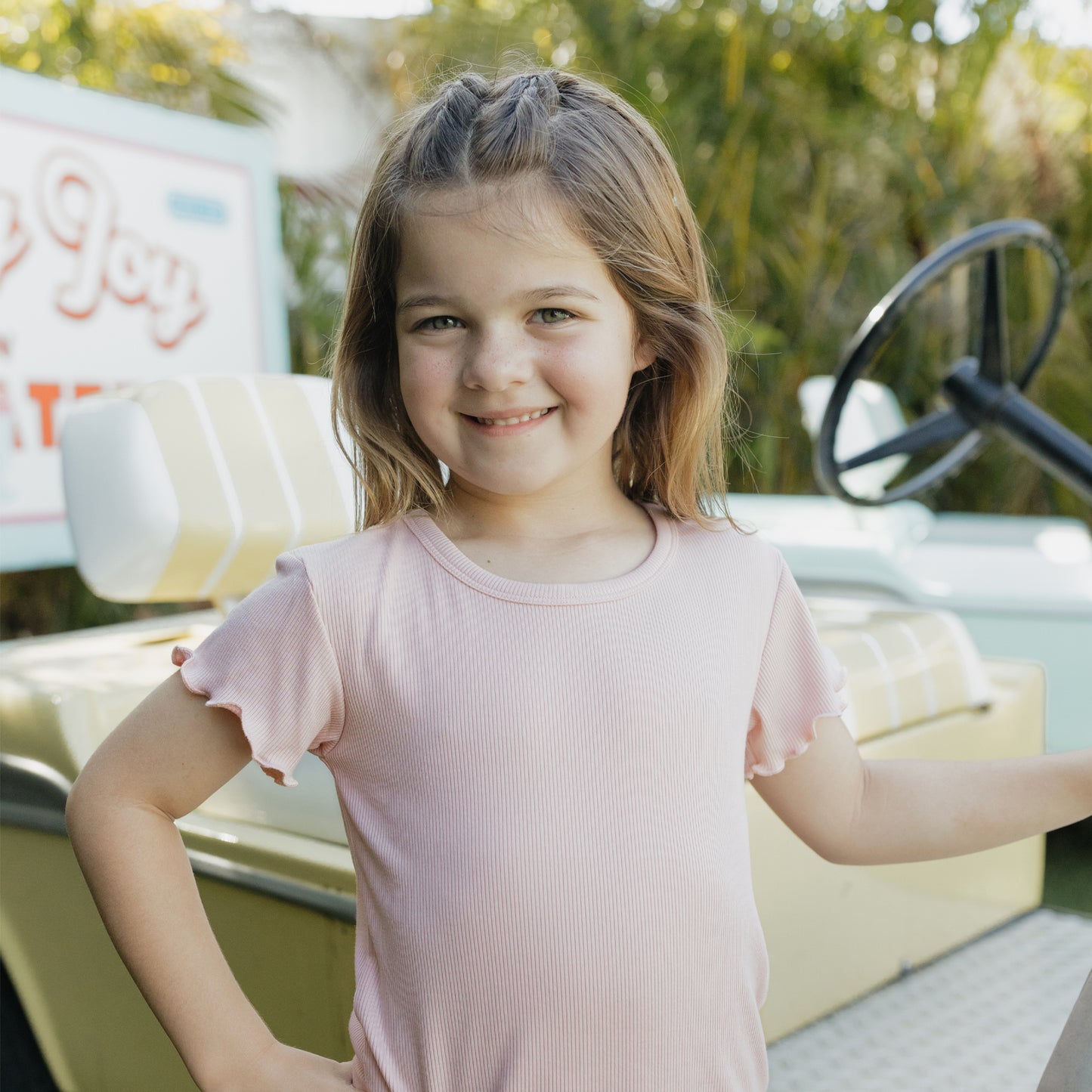 Young girl in a pink shirt standing in front of a vintage-style vehicle with a blurred background
