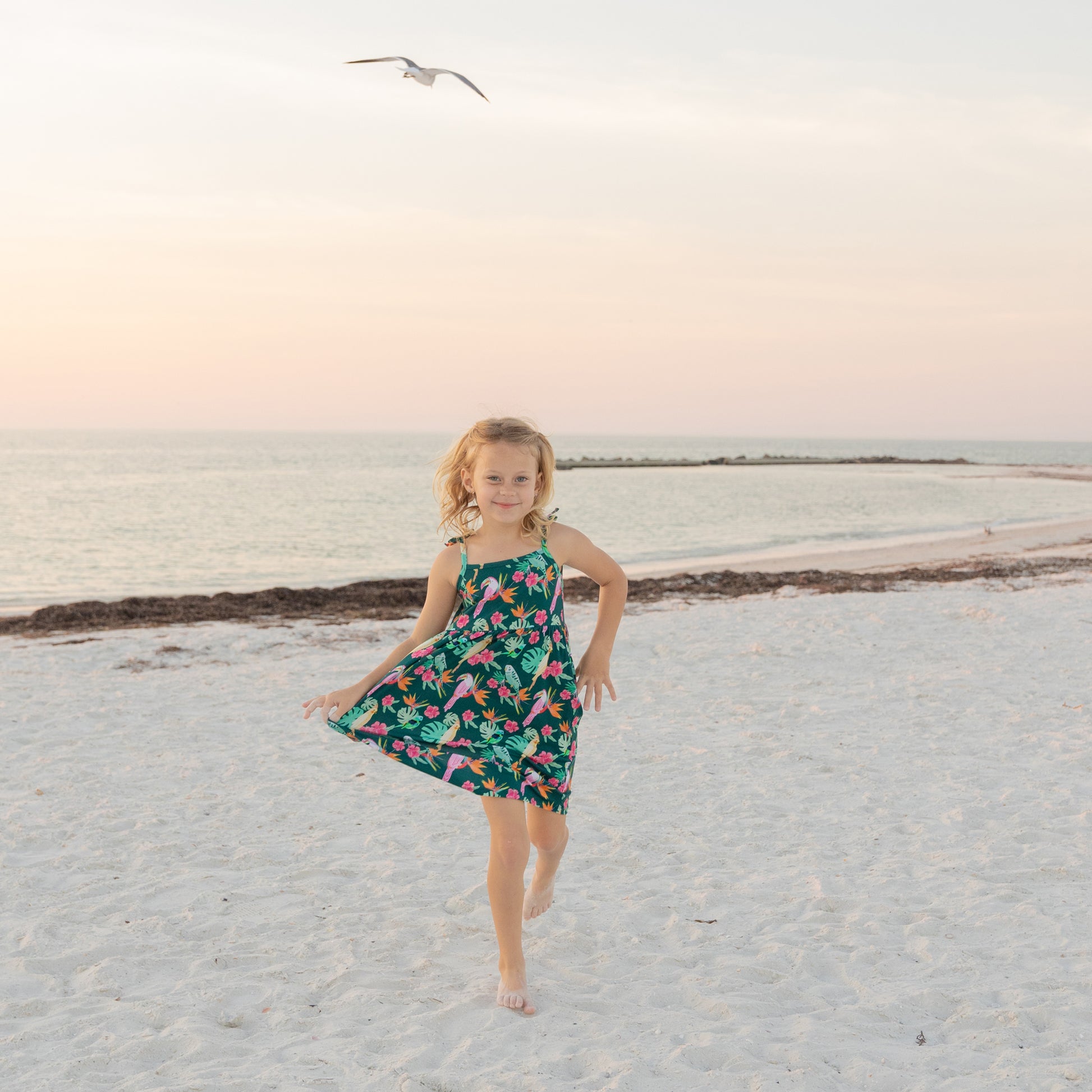 A young girl standing on a beach at sunset wearing the Isla Botánica bamboo sundress, showcasing the breezy, summer fit.