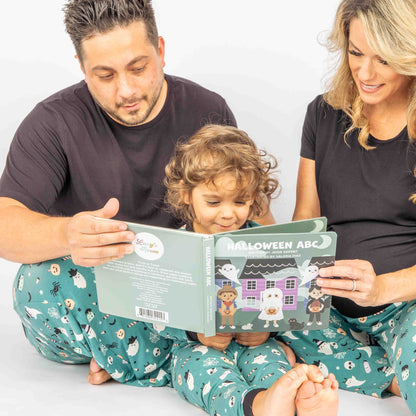 Mom and Dad holding Halloween ABCs book for little girl to read from while sitting on the ground. They are all wearing bamboo pajamas in the "Boo Crew" Print
