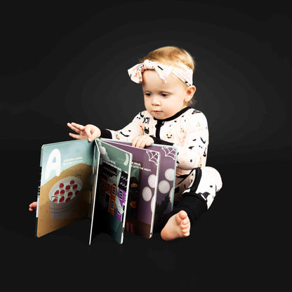 Little girl sitting while holding halloween abc board book modeling the Sweet and Spooky halloween print