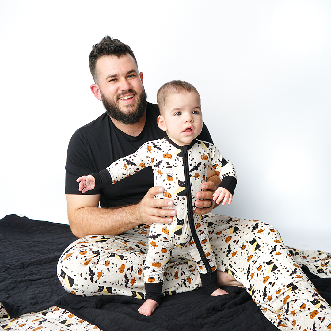 dad sits on the "spooky cute beige" quilt and wears his "spooky cute beige" relaxed pajama pants. he hold his son up, who is also wearing "spooky cute beige" in the convertible. the "spooky cute beige" print is a halloween themed pattern that contains cute versions go ghost, skeletons, black cats, candy treats, bats, and pumpkins. 