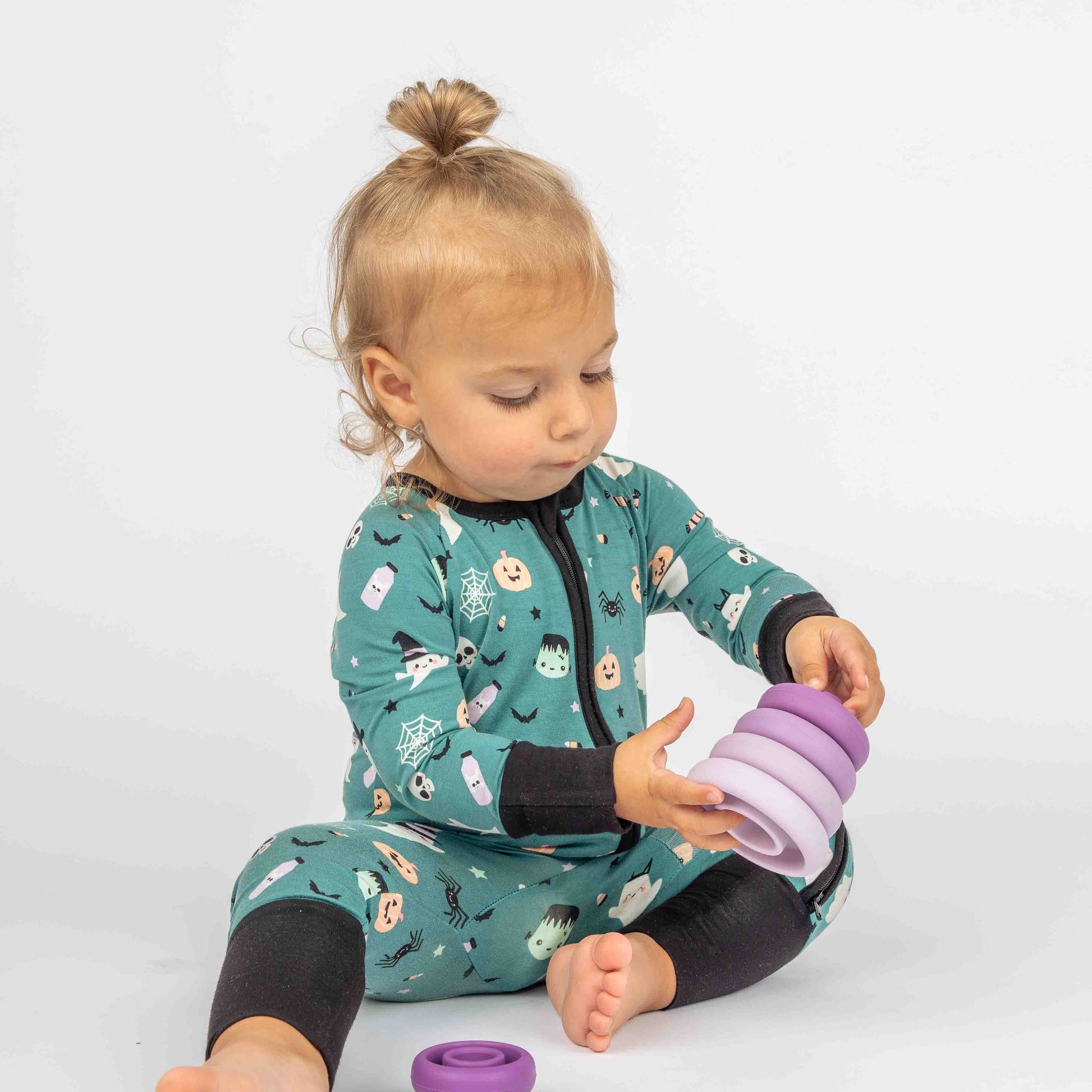 Baby sitting on ground playing with purple stacking silicone toy while wearing "Boo Crew" our halloween print featuring glow in the dark white ghosts and stars, pumpkins, spiders, webs, candy, Frankenstein, and more!