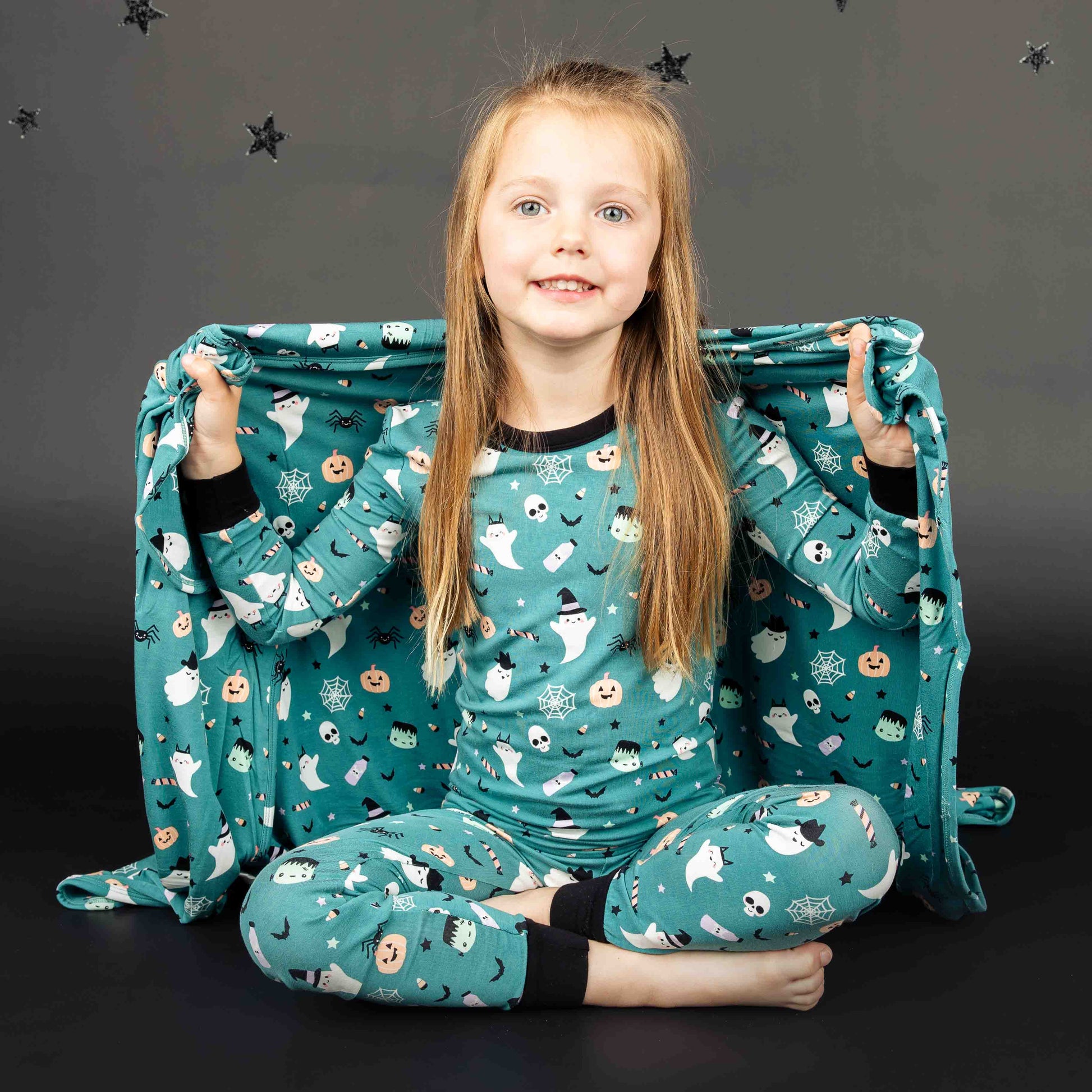 Girl sitting while smiling at the camera, holding the baby blanket behind her and wearing the toddler long-sleeve pajama set. Both products are in the "Boo Crew" halloween print featuring glow in the dark white ghosts and stars, pumpkins, spiders, webs, candy, Frankenstein, and more! 