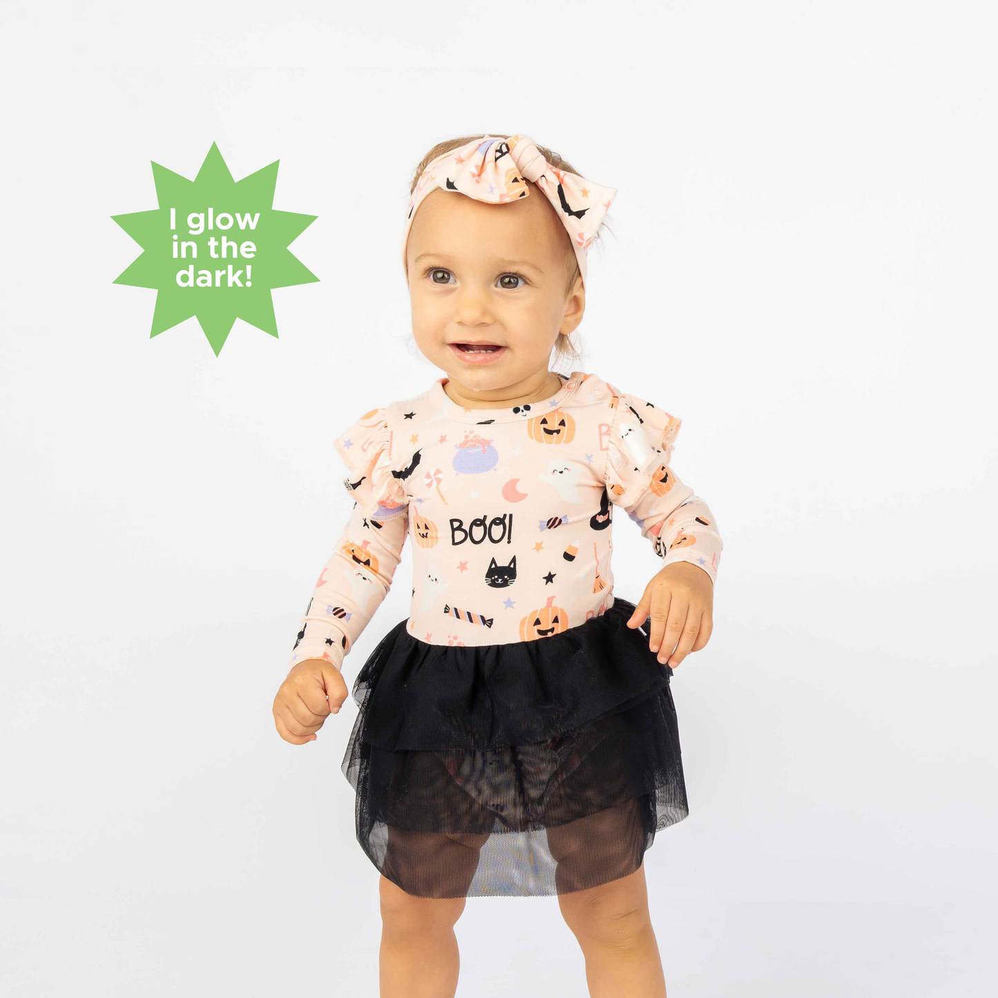 Baby standing on white backdrop modeling the longsleeve skirted onesie and bow in our Sweet and Spooky pink halloween Print featuring glow in the dark patterns, pumpkins, candy, bats and more!