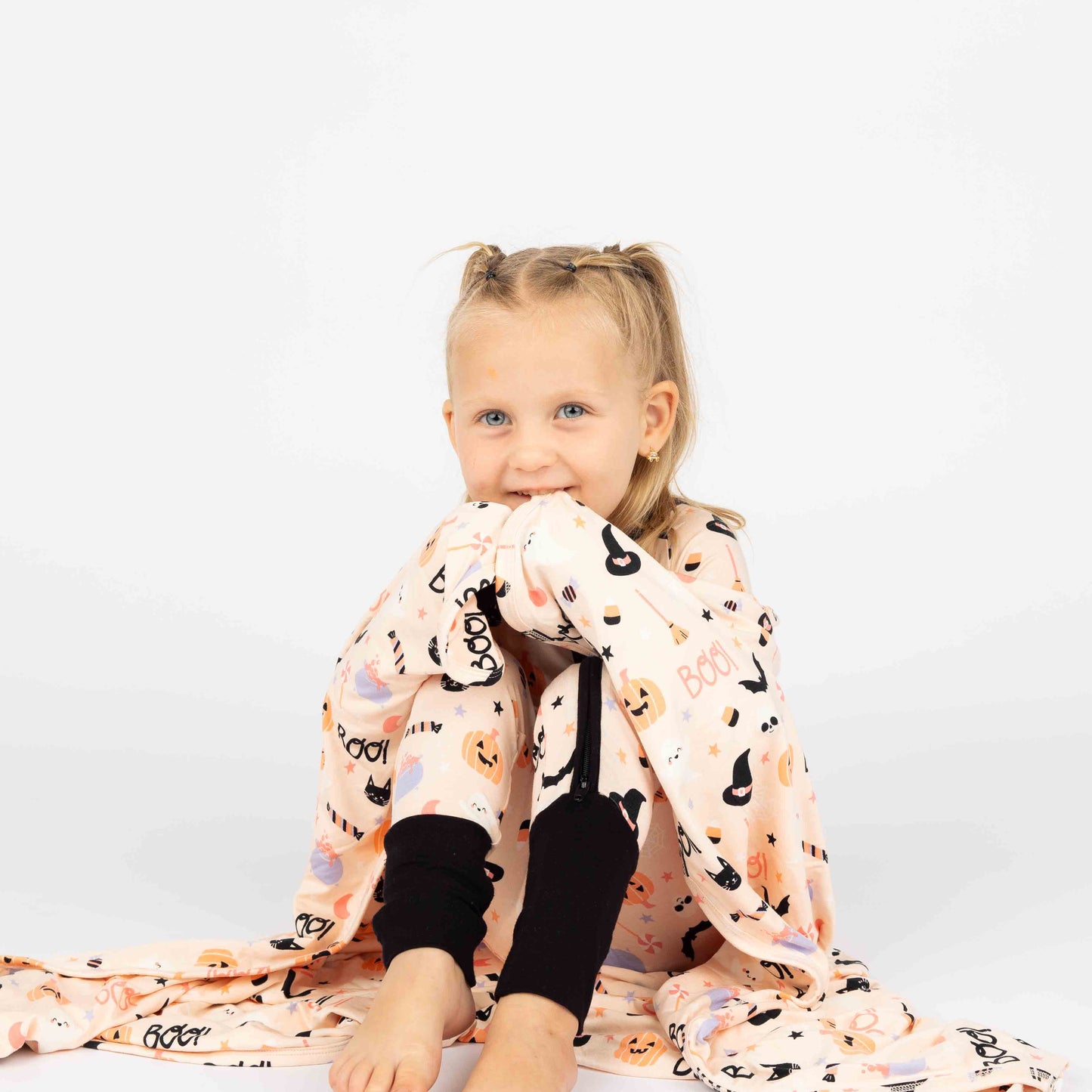 Girl sitting wrapped up in our bamboo halloween blanket and wearing our convertible pajama. Both products are in  our Sweet and Spooky pink halloween Print featuring glow in the dark patterns, pumpkins, candy, bats and more!