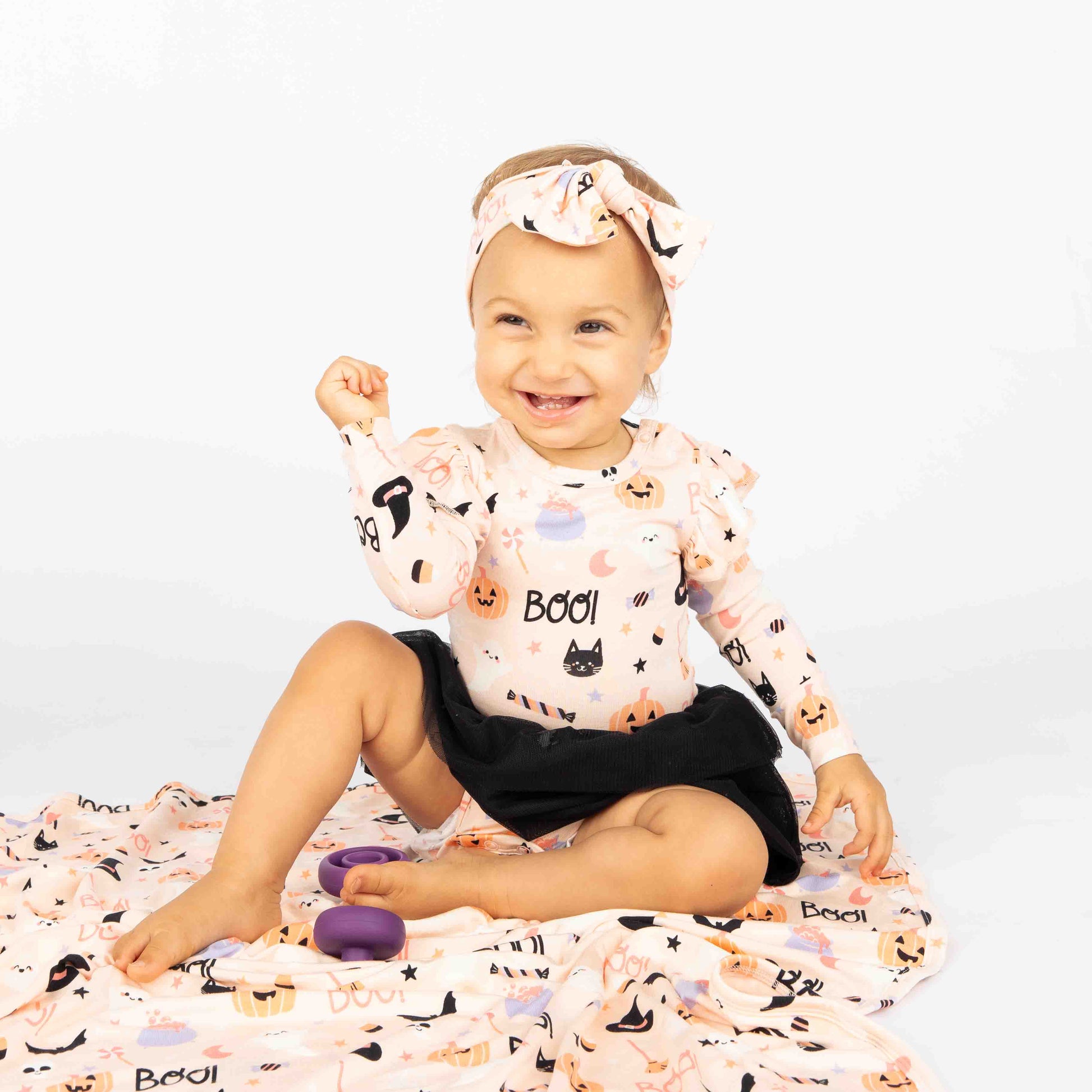 Baby wearing our skirted onesie style sitting on top of the baby bamboo blanket. Both products she is modeling are in  our Sweet and Spooky pink halloween Print featuring glow in the dark patterns, pumpkins, candy, bats and more!