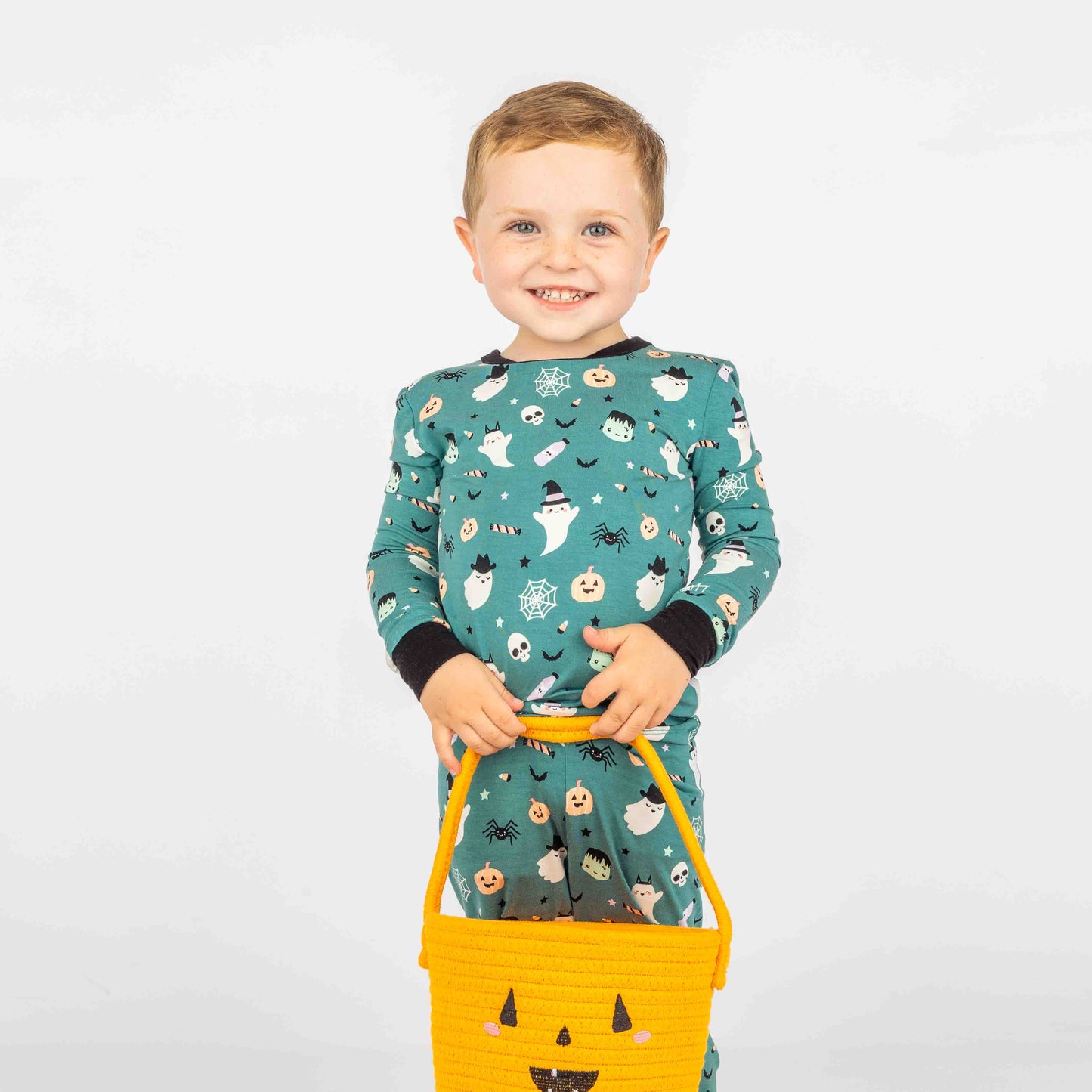 Kid standing in front of white background holding an orange halloween pumpkin rope basket and modeling "Boo Crew" our halloween print featuring glow in the dark white ghosts and stars, pumpkins, spiders, webs, candy, Frankenstein, and more! 