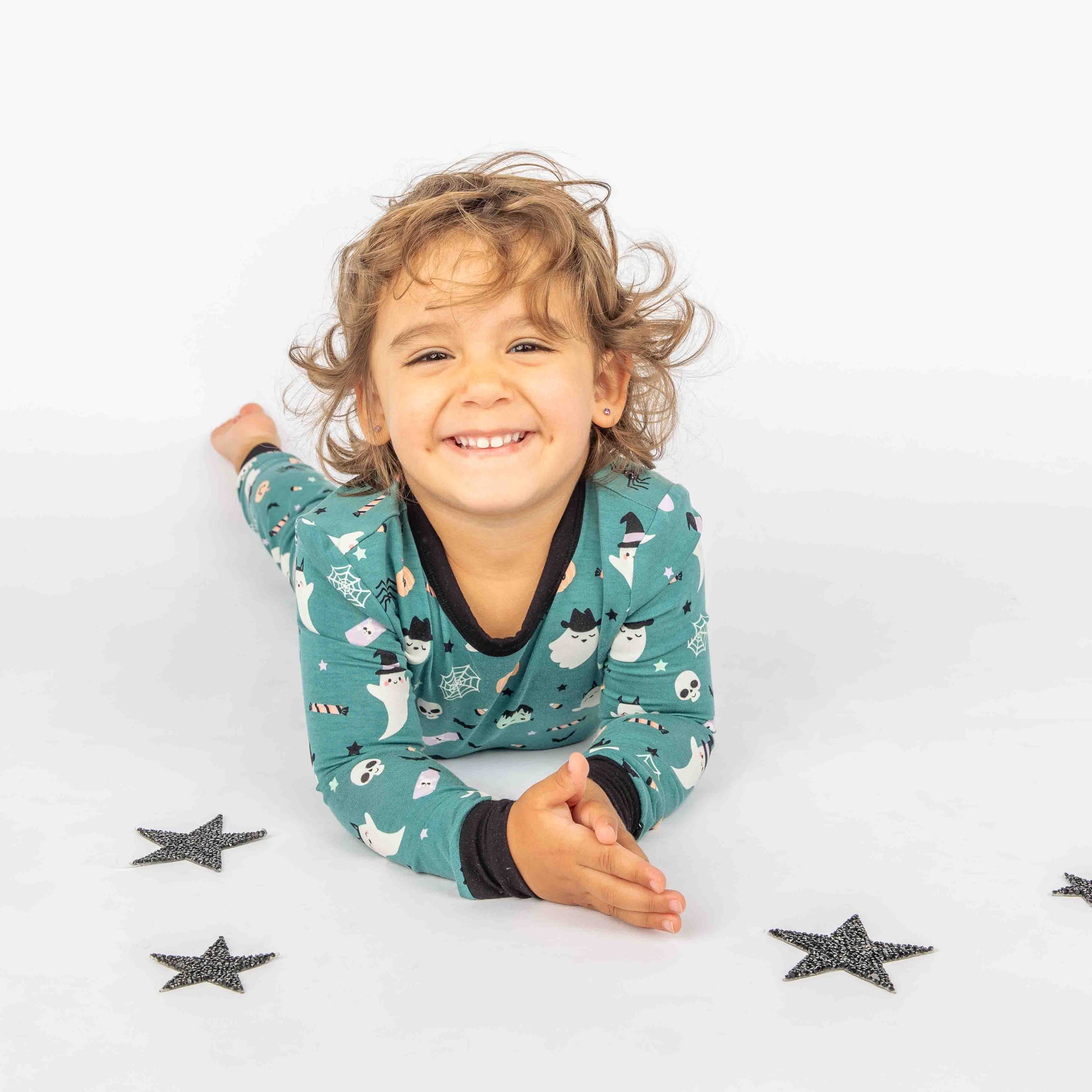 Kid smiling at camera while laying on their belly modeling our "Boo Crew" our halloween print featuring glow in the dark white ghosts and stars, pumpkins, spiders, webs, candy, Frankenstein, and more!