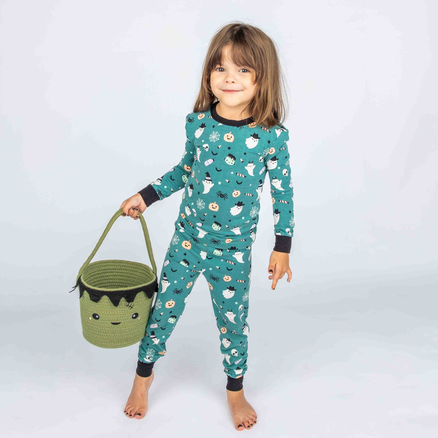 Kid holding green Frankenstein basket while standing in the kids pajama set on our "Boo Crew" our halloween print featuring glow in the dark white ghosts and stars, pumpkins, spiders, webs, candy, Frankenstein, and more!