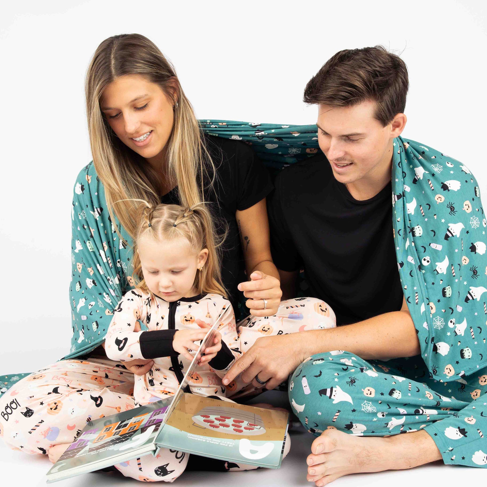 Family reading while cuddling under a twin size bamboo blanket in the "Boo Crew" our halloween print featuring glow in the dark white ghosts and stars, pumpkins, spiders, webs, candy, Frankenstein, and more!