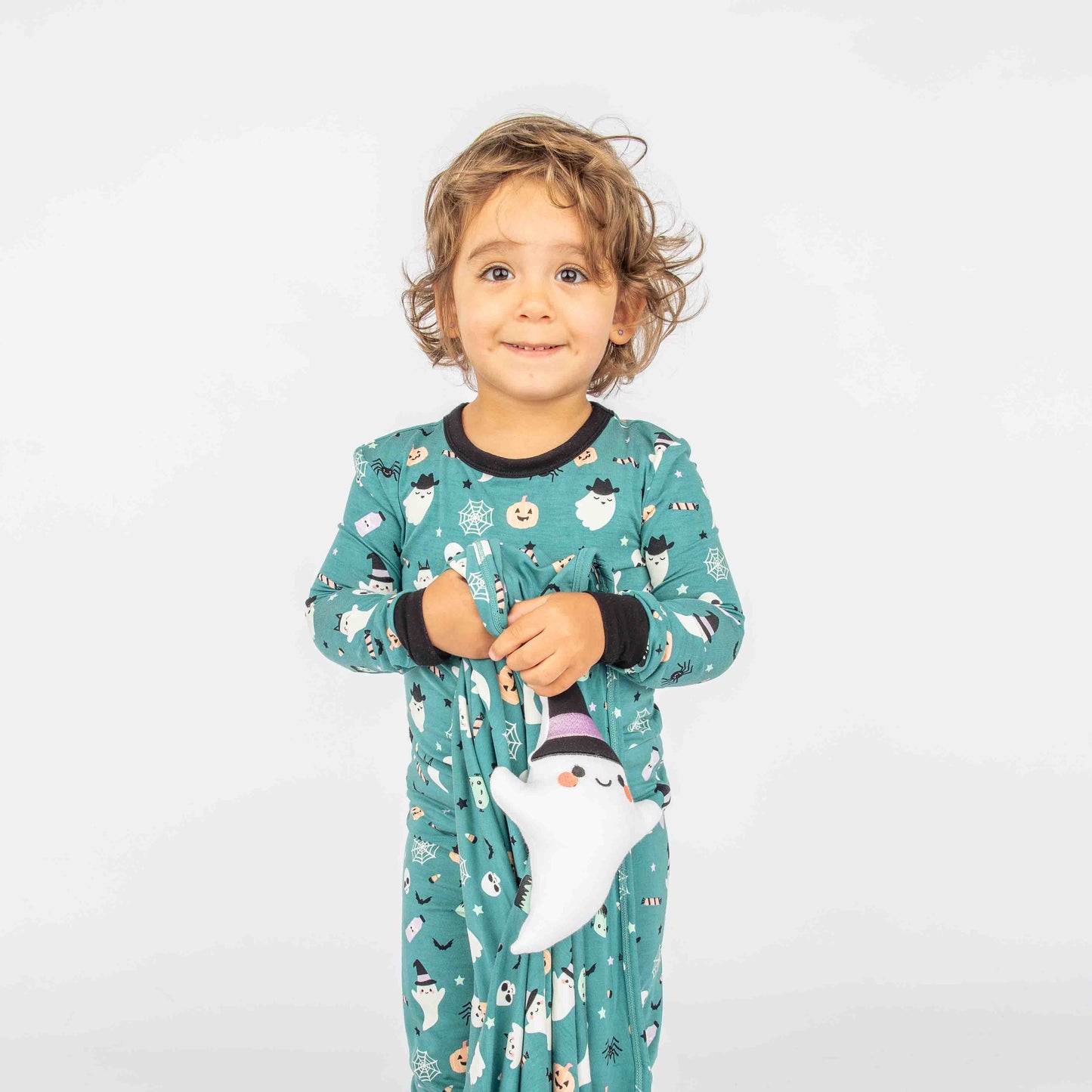 Little Kid Standing and smiling while holding a halloween ghost lovey. She is wearing "Boo Crew" halloween print featuring glow in the dark white ghosts and stars, pumpkins, spiders, webs, candy, Frankenstein, and more! 