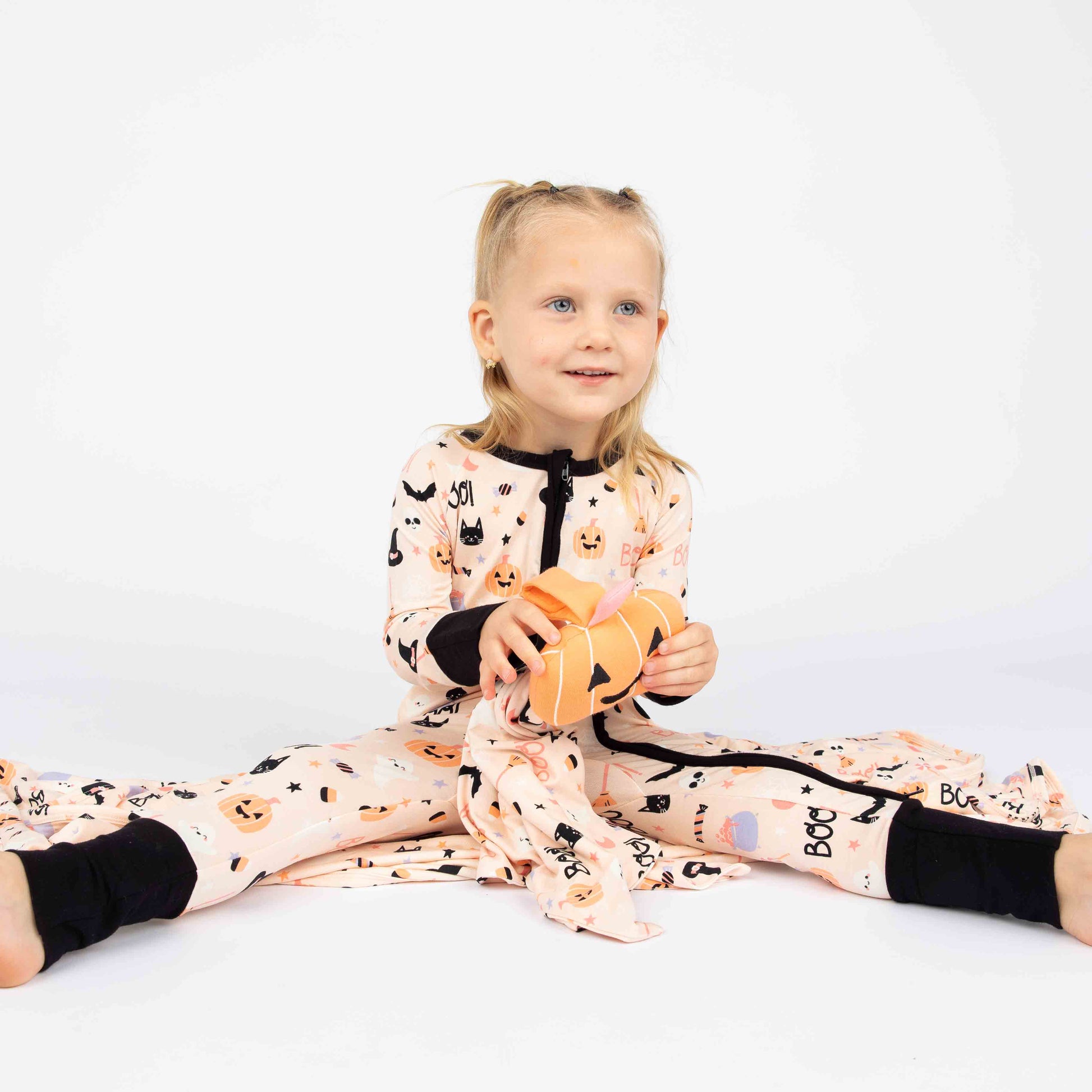 girl sitting on white background while holding a pumpkin lovey and wearing our Sweet and Spooky pink halloween Print featuring glow in the dark patterns, pumpkins, candy, bats and more!