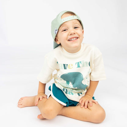 Little boy sitting with the Happiness Comes in Waves hat on backwards while wearing the Emerson and Friends Save the Manatees Cotton Tee with terry cloth shorts. 